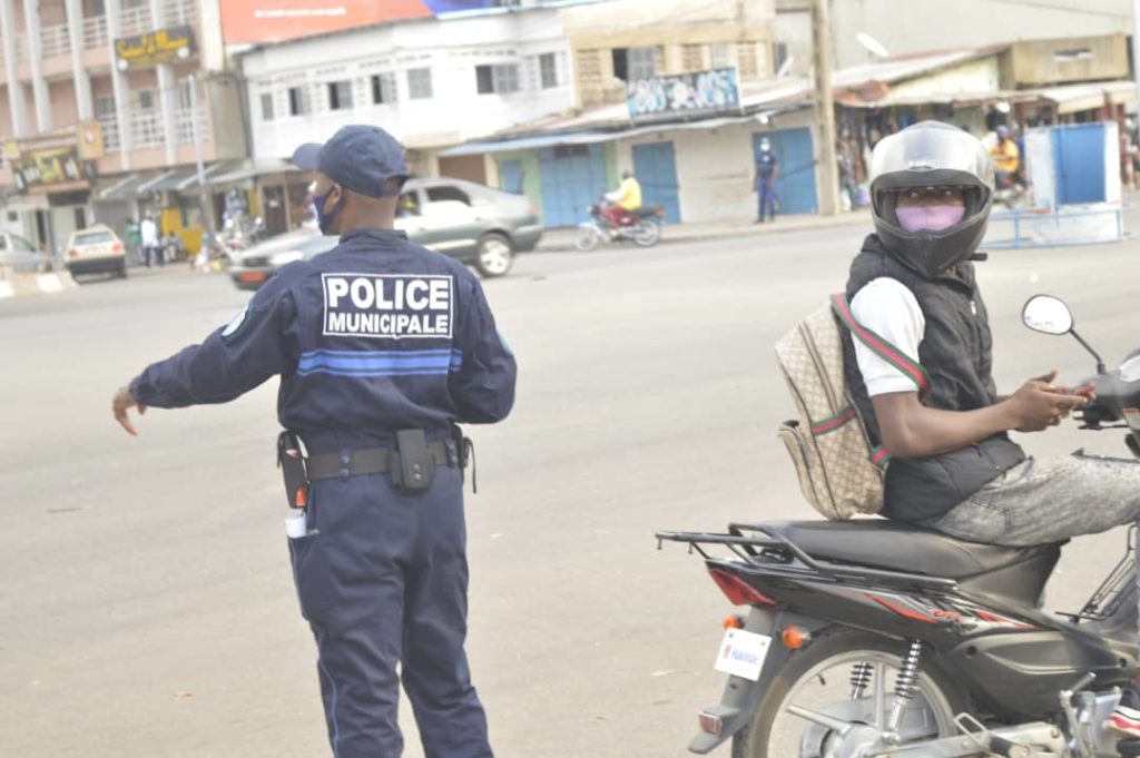 Un agent de police municipale en uniforme dirige la circulation tandis qu’un motard masqué attend sur sa moto.