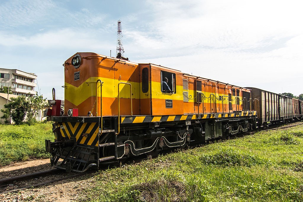 Locomotive orange et jaune tractant des wagons marchandise sur une voie ferrée environnée de verdure et de bâtiments en arrière-plan