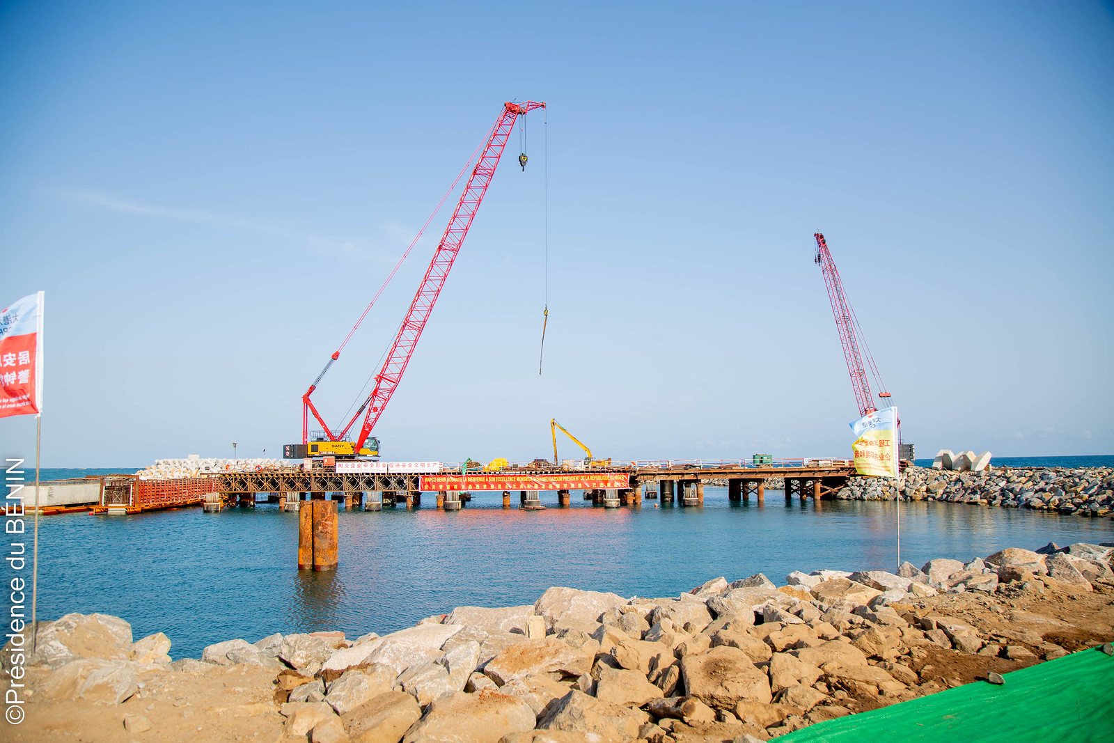 Deux grues rouges installées sur une structure en construction au-dessus de l'eau avec des rochers au premier plan