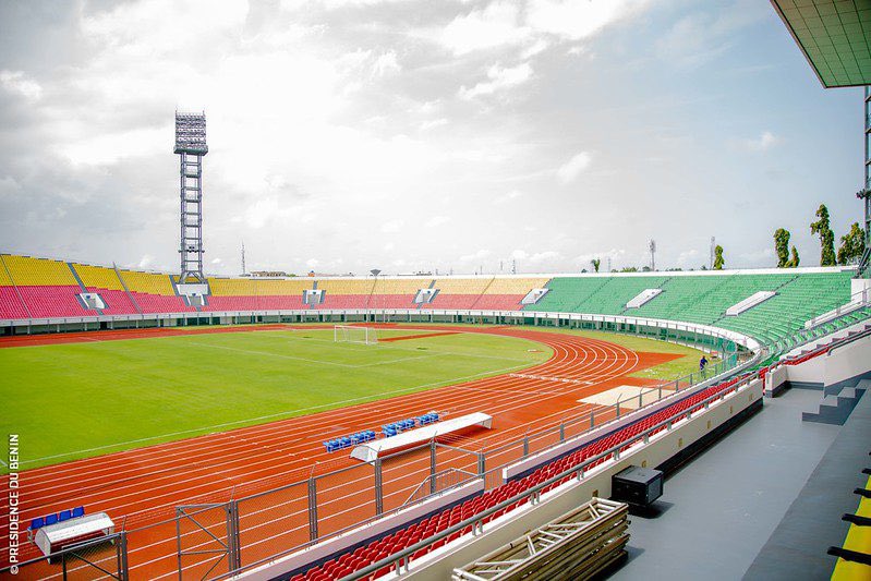 Stade avec pistes d'athlétisme et tribunes colorées en rouge, jaune et vert sous un ciel partiellement nuageux