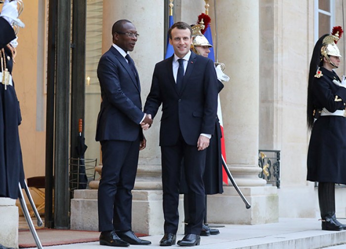 Deux hommes en costume sombre se serrent la main devant un bâtiment avec des gardes en uniforme et casque à plumes rouges.