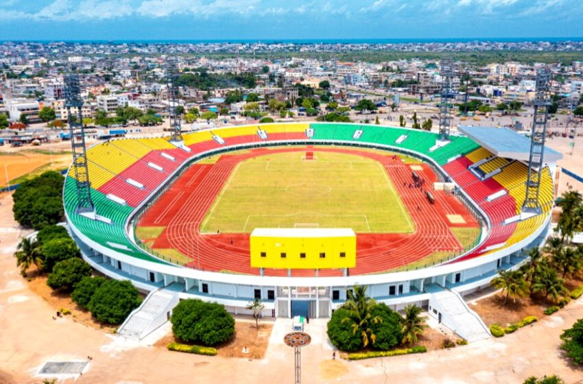 Stade de football avec piste d’athlétisme et tribunes colorées vu en contre-plongée aérienne avec ville en arrière-plan