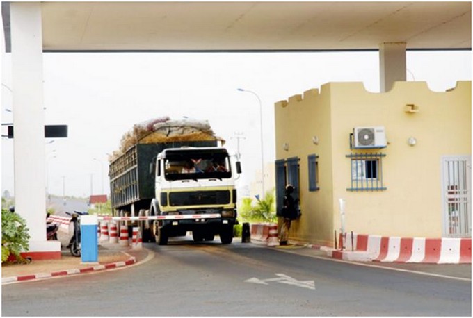 Camion chargé passant sous un poste de péage avec une barrière et un bâtiment de contrôle à côté