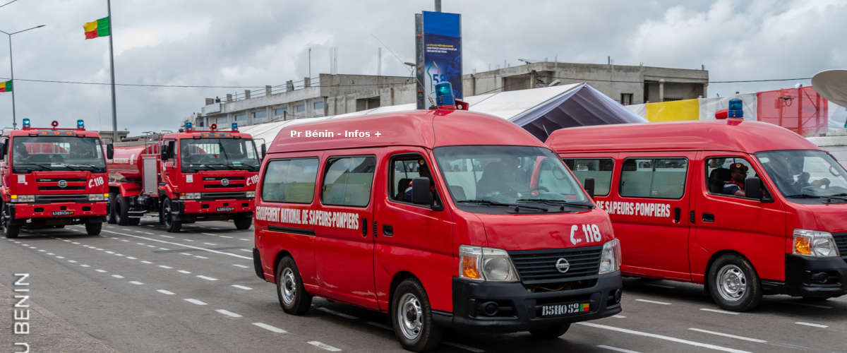 Plusieurs véhicules rouges de sapeurs-pompiers alignés sur une route avec un drapeau en arrière-plan sous un ciel nuageux