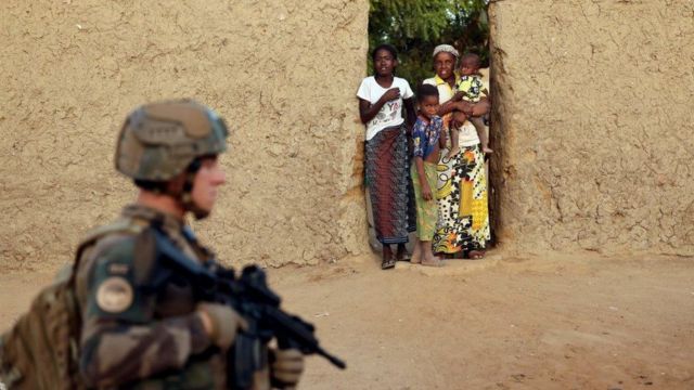Soldat en uniforme armé en premier plan, femmes et enfants debout dans l’embrasure d’une porte en terre battue en arrière-plan