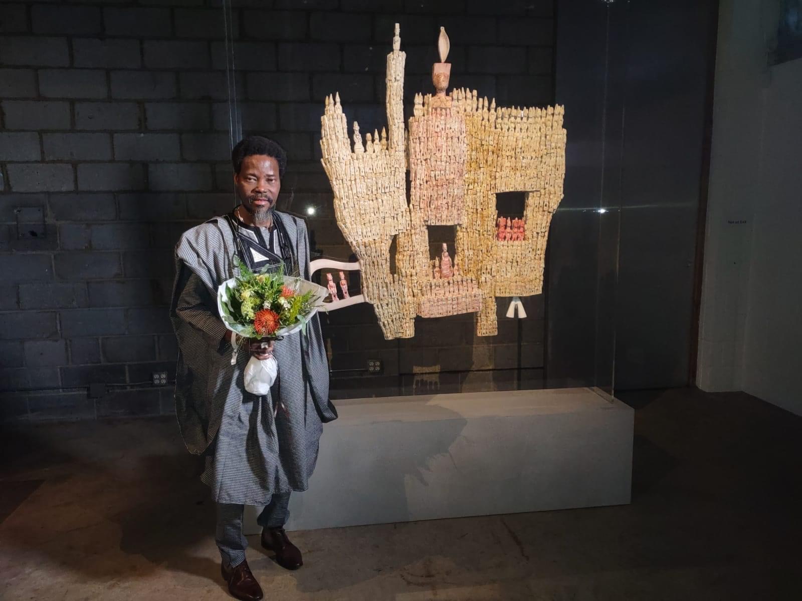 Un homme en tenue traditionnelle tient un bouquet près d'une sculpture en forme de main ornée de petites figurines en exposition
