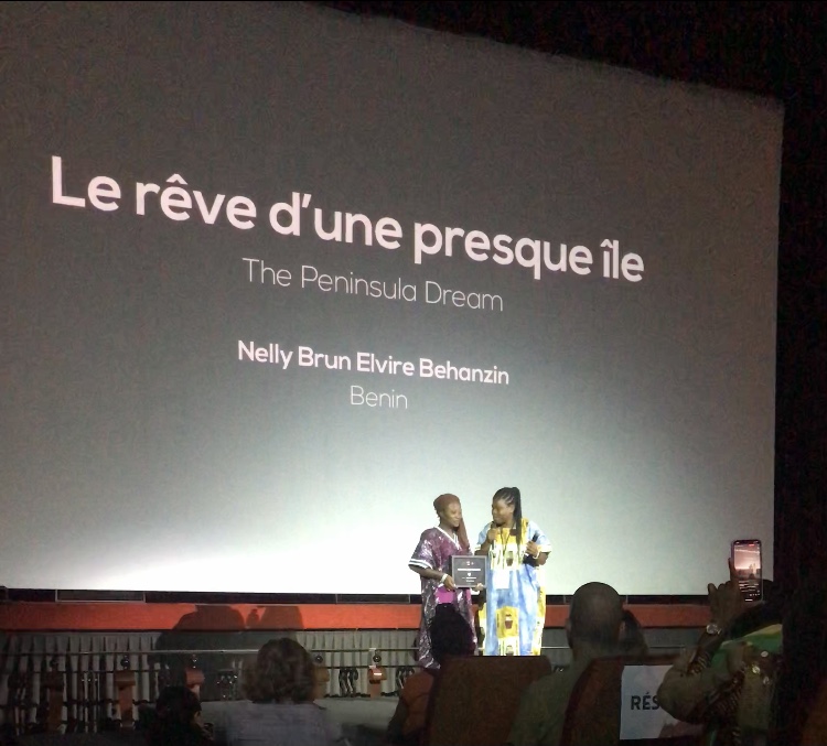 Deux personnes sur scène devant un grand écran portant le titre d’un film et un nom avec un public assis au premier plan