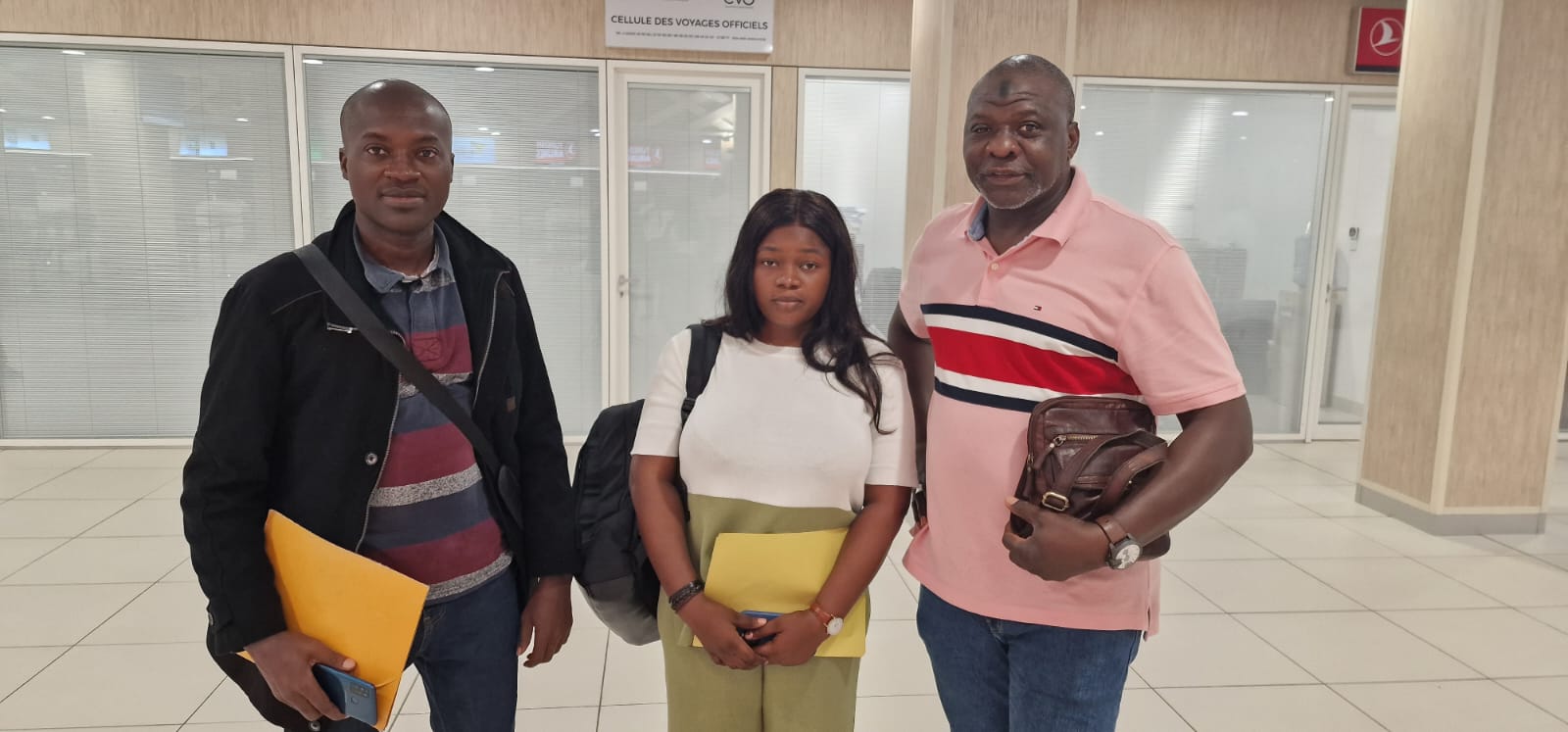 Trois personnes souriantes posent dans un hall intérieur avec des dossiers jaunes à la main et un sac à dos noir visible