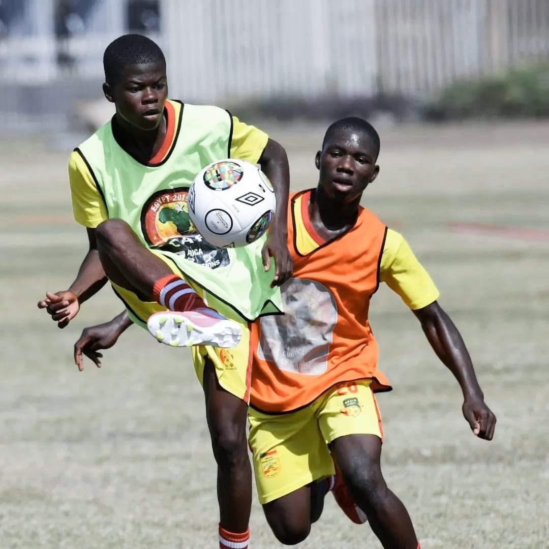 Deux jeunes joueurs en maillots jaunes et chasubles verte et orange disputent un ballon en plein air sur un terrain de foot