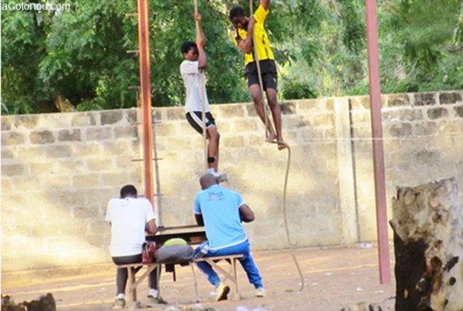 Deux adolescents grimpent à la corde tandis que deux adultes assis à une table semblent noter ou observer l'épreuve sportive