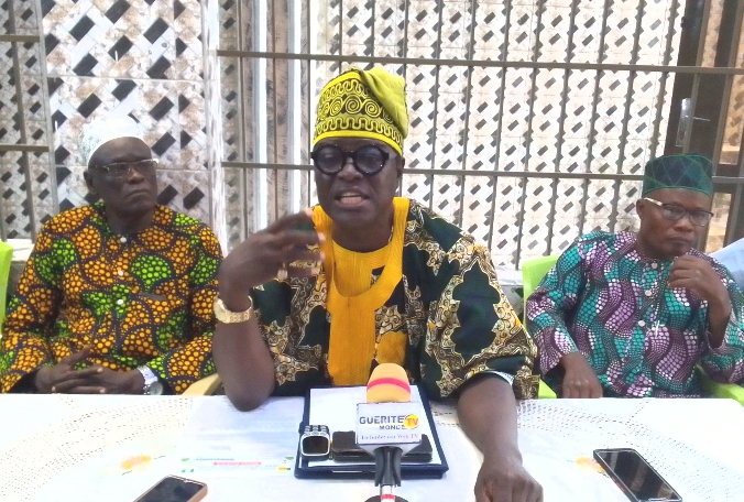 Trois hommes en tenue traditionnelle colorée sont assis derrière une table avec un micro et plusieurs téléphones posés dessus
