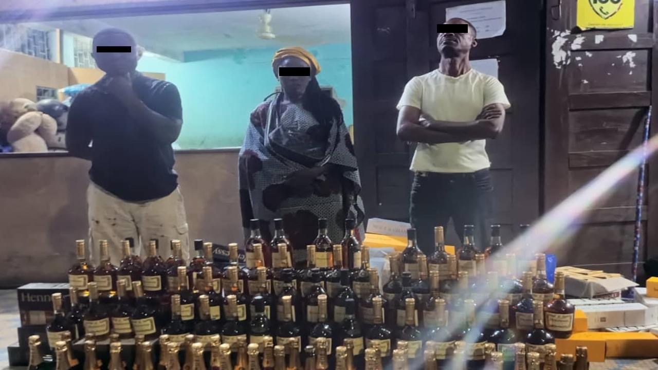 Trois individus masqués debout derrière une grande quantité de bouteilles d'alcool empilées sur une table devant une porte ouverte