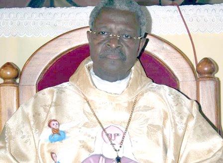 Homme âgé avec lunettes vêtu d’une chasuble dorée assis sur un siège en bois avec coussin rouge