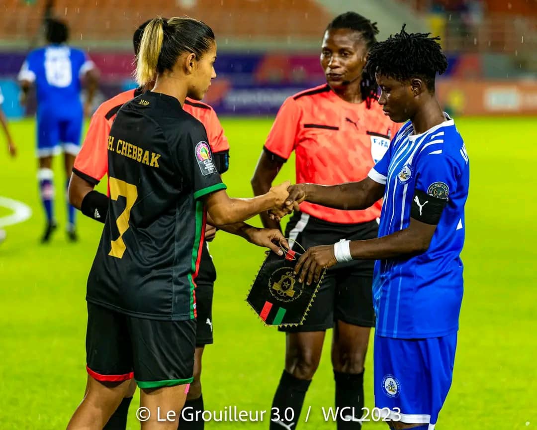 Deux joueuses échangent un objet avant le début du match sous le regard d'une arbitre en tenue orange sur le terrain de football humide