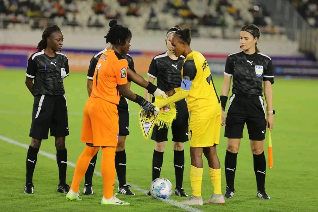 Deux capitaines d’équipes féminines de football échangent un fanion devant trois arbitres sur un terrain de stade quasi vide