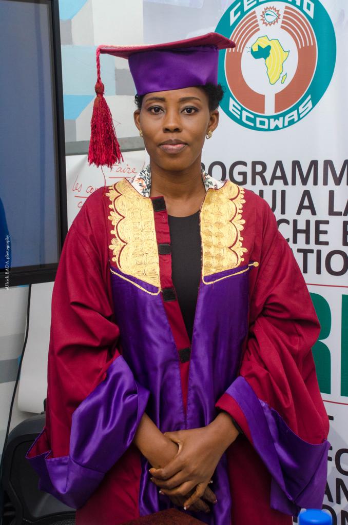 Jeune femme en tenue de doctorat violette et rouge posant devant un panneau avec logo Afrique et texte programmatique
