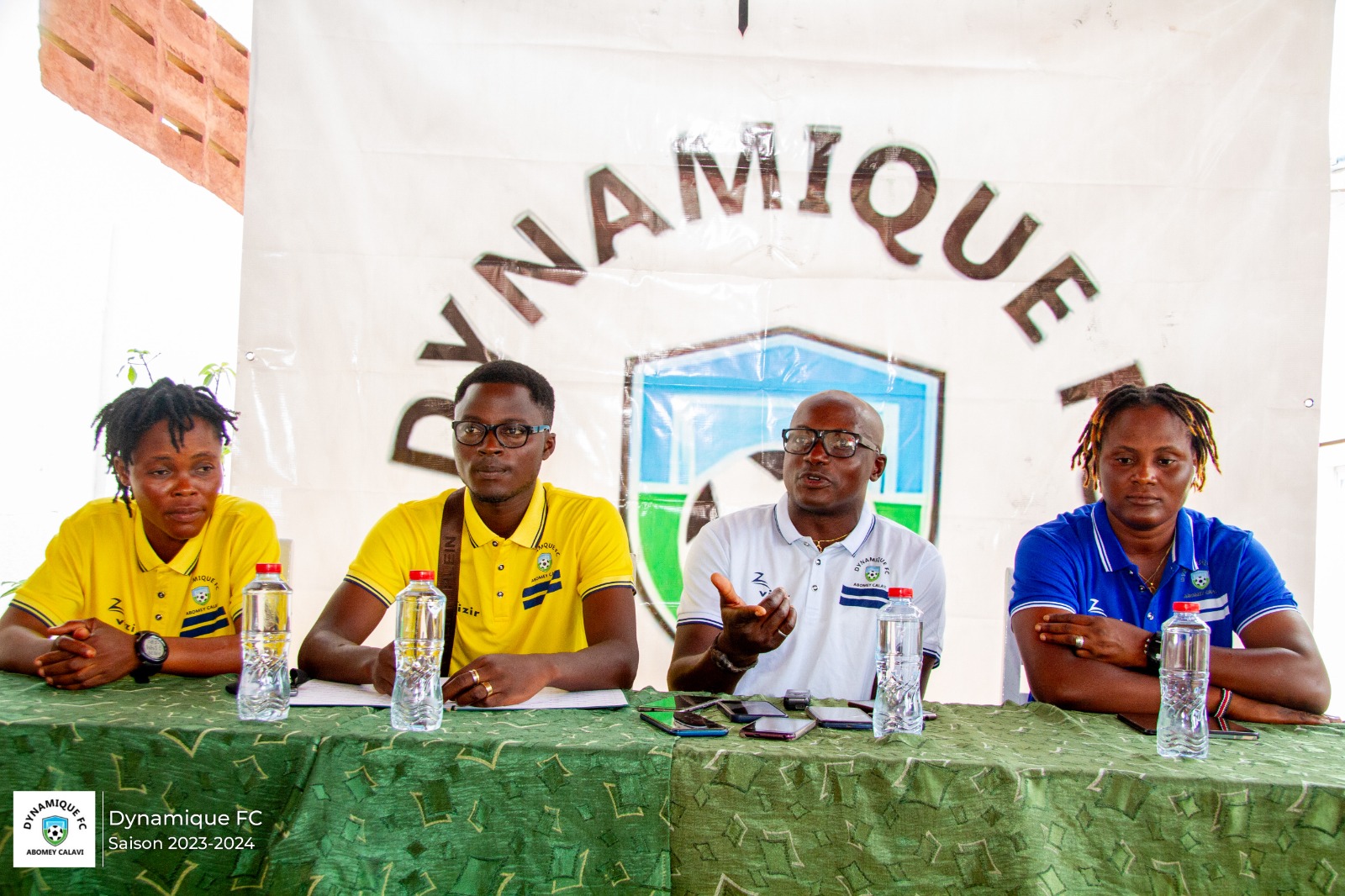 Quatre personnes assises en rang derrière une table verte avec bouteilles d’eau et un grand logo sportif en arrière-plan