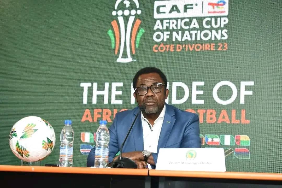 Homme assis derrière une table avec micro, devant un écran affichant le logo de la CAN Côte d’Ivoire 2023 et une balle de football posé à côté de bouteilles d’e