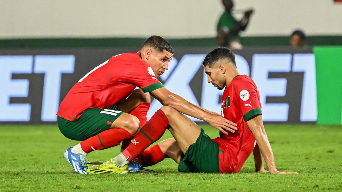 Un joueur en rouge aide un coéquipier assis au sol lors d’un match de football sur pelouse verte