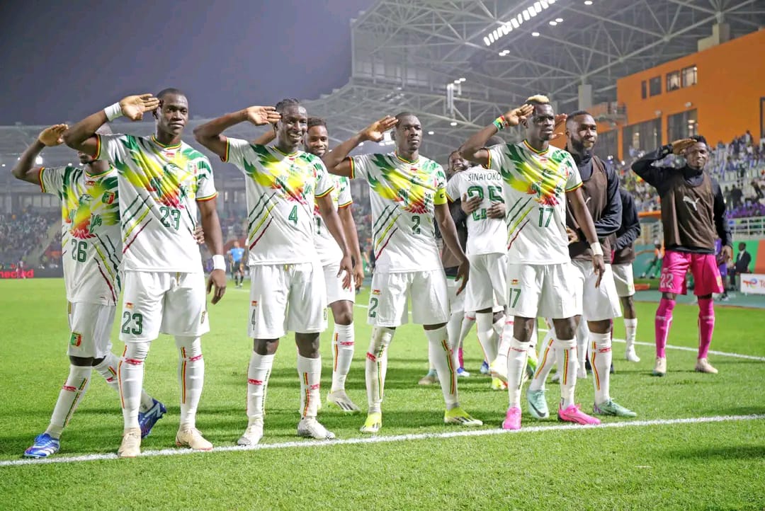 Des joueurs de football en tenue blanche et colorée saluent en levant la main au bord du terrain sous un stade éclairé