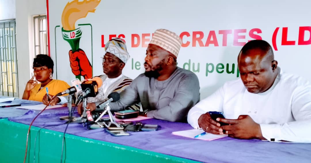 Quatre personnes assises à une table devant une banderole avec un logo et le texte partiel « LES DÉMOCRATES » discutent ou écoutent
