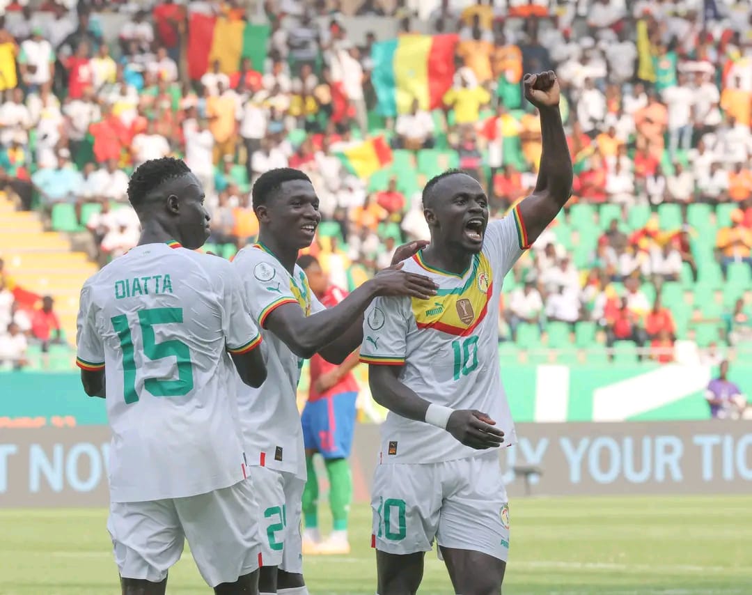 Trois joueurs de football en maillot blanc célèbrent un but sur le terrain devant des gradins colorés et un drapeau en arrière-plan
