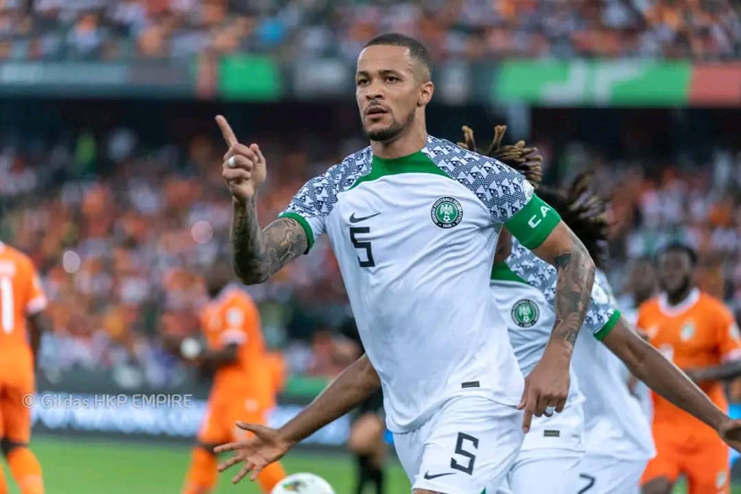 Un joueur en maillot blanc lève le doigt lors d’un match de football contre une équipe en maillot orange dans un stade rempli