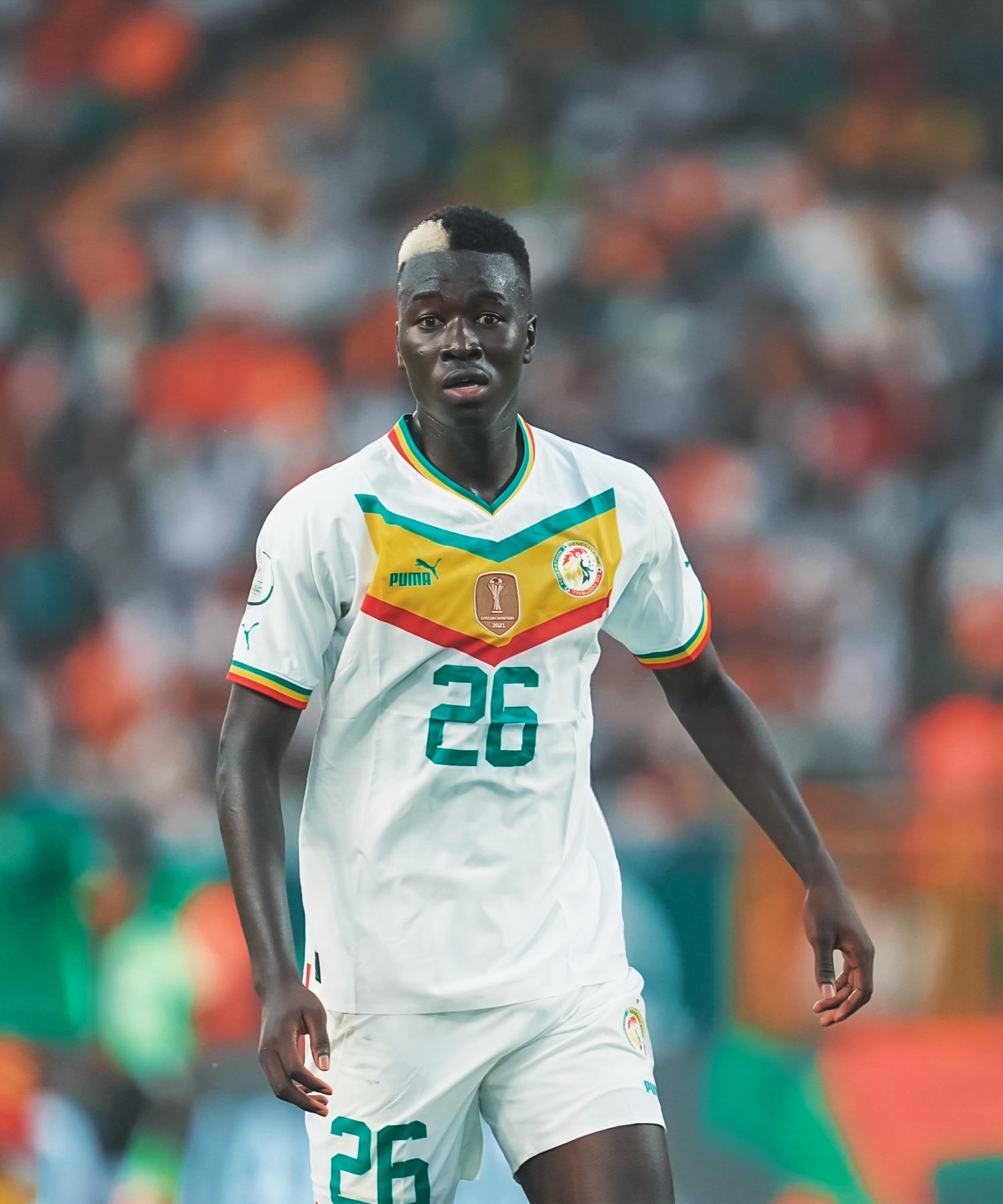 Un joueur de football en maillot blanc avec le numéro 26 sur un terrain avec des spectateurs flous en arrière-plan