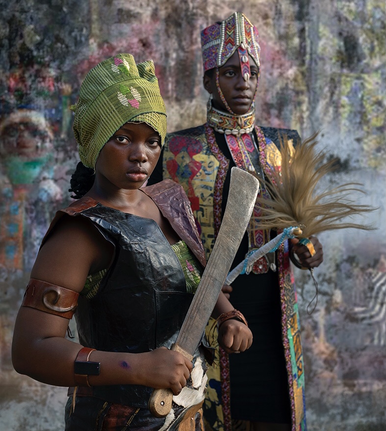 Deux femmes en costumes traditionnels, l’une brandit un grand couteau, l’autre tient un objet orné de plumes devant un mur coloré