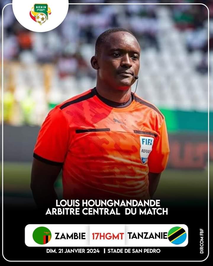 Un arbitre en tenue orange avec un badge FIFA est au premier plan dans un stade aux gradins flous en arrière-plan