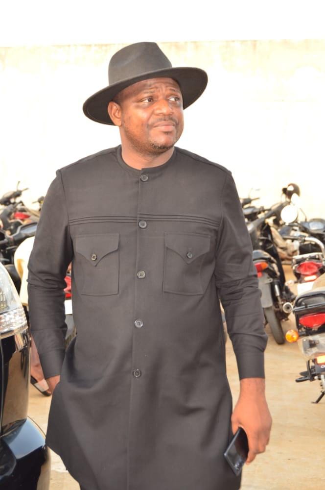 Homme debout portant un manteau noir et un chapeau noir, tenant un téléphone portable, avec plusieurs motos en arrière-plan