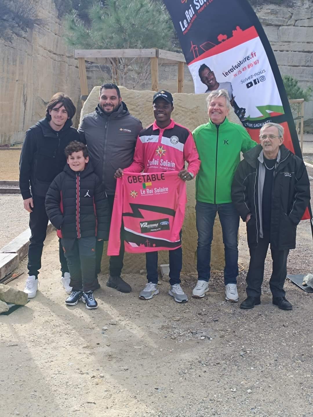 Six hommes posent debout sur un terrain avec un maillot rose tenu par l’homme central devant un panneau publicitaire.