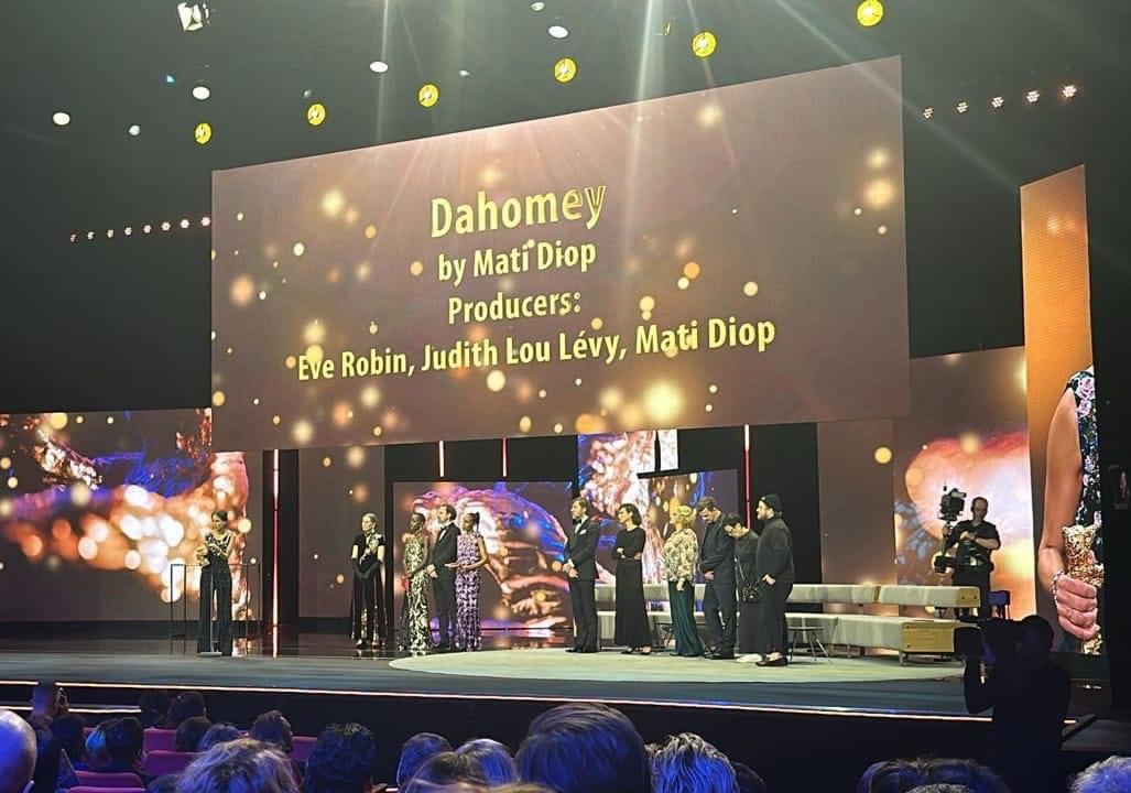 Personnes sur scène recevant un prix avec un grand écran affichant le titre et producteurs du film Dahomey en arrière-plan