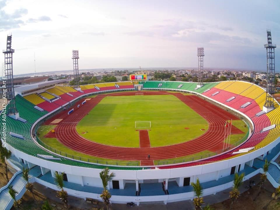 Stade circulaire avec piste d’athlétisme et pelouse verte entourée de gradins rouges, jaunes et verts sous ciel partiellement nuageux