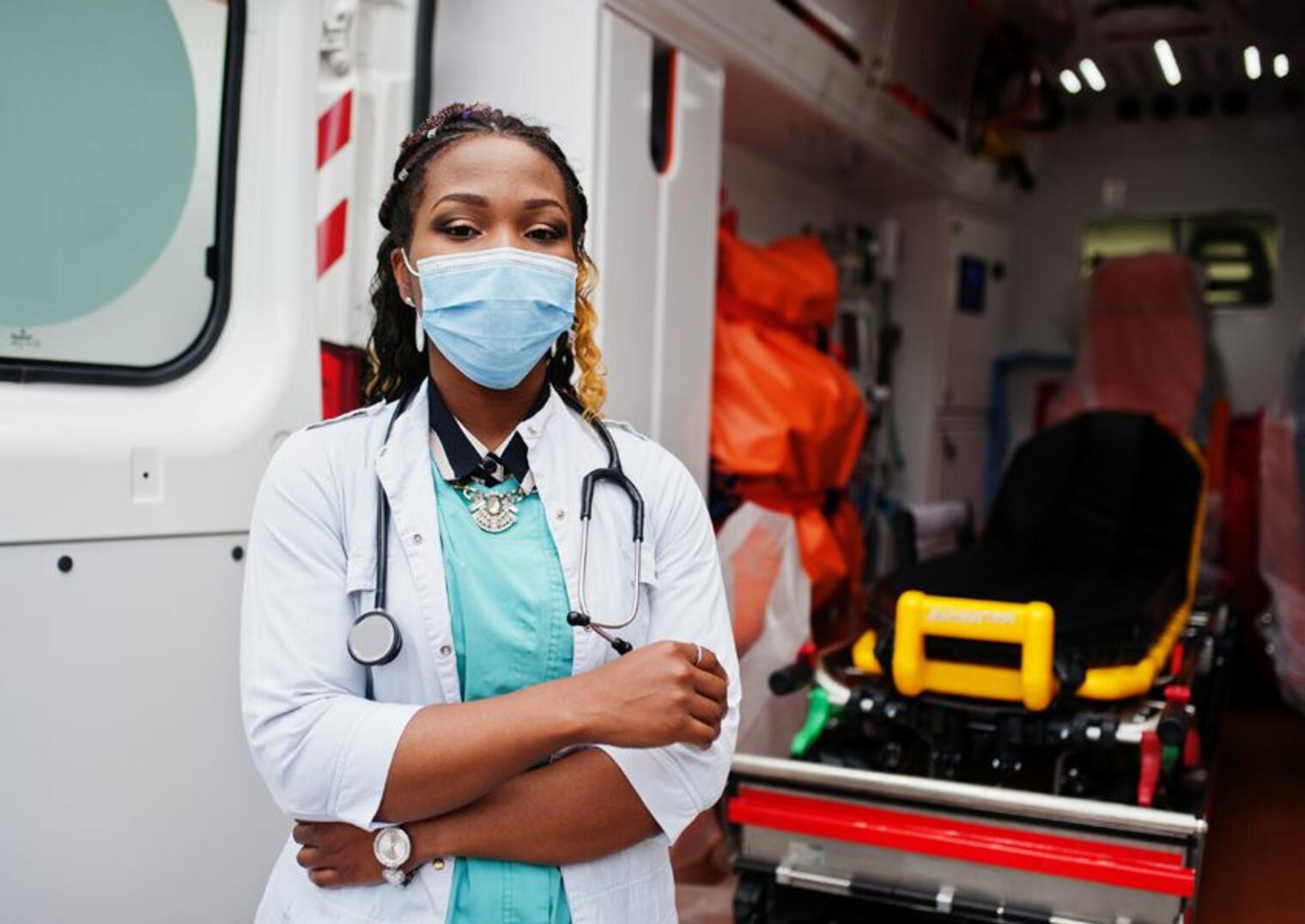 Jeune professionnelle de santé portant une blouse blanche et un masque, debout devant une ambulance avec un brancard visible à l’intérieur