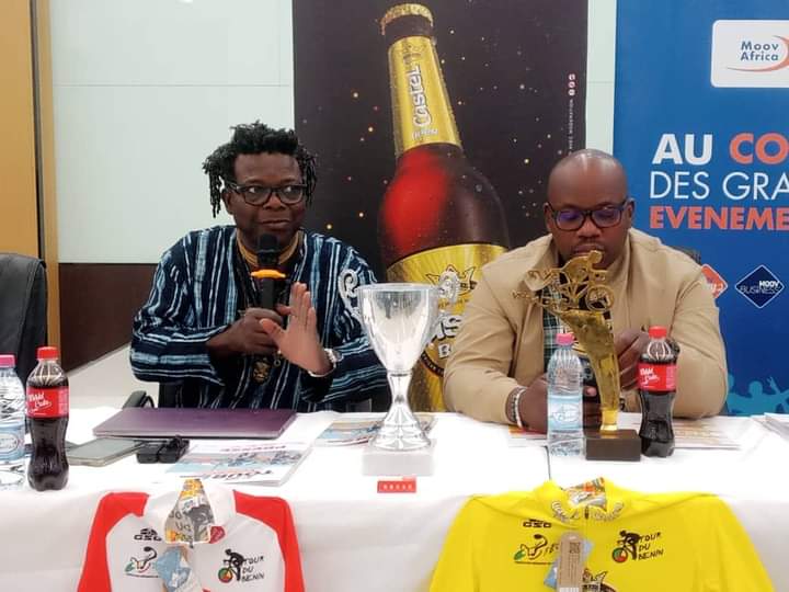 Deux hommes assis à une table avec des trophées, des maillots de cyclisme et des bouteilles de boisson devant eux
