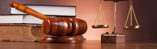 Un marteau de juge posé sur un socle en bois avec une balance de justice en arrière-plan sur une surface marron