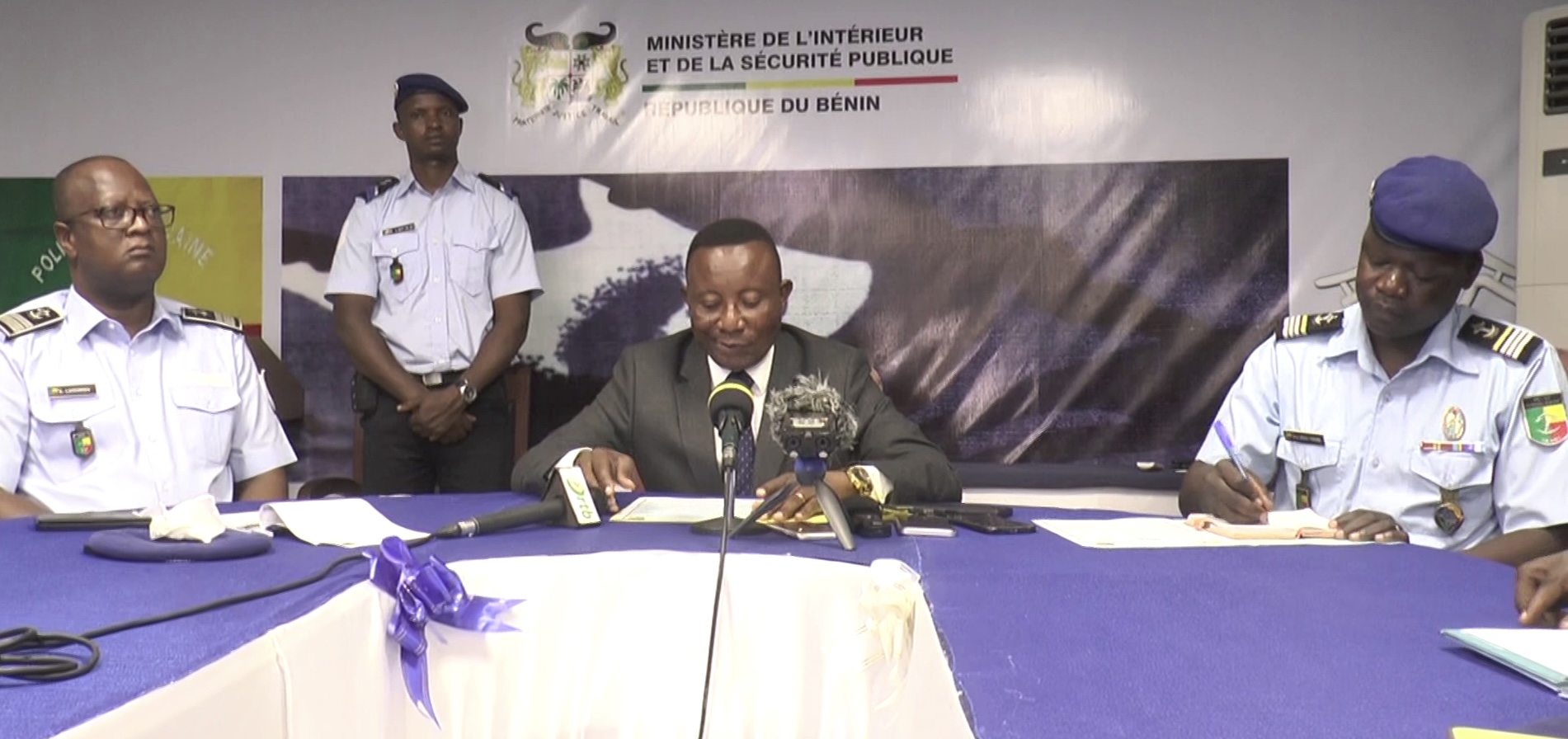 Quatre hommes en uniformes et costume assis à une table avec microphones et documents devant un panneau du ministère de l’Intérieur et de la Sécurité publique