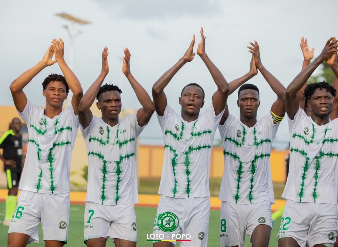Six joueurs de football en maillot blanc à motifs verts applaudissent côte à côte sur un terrain de jeu.