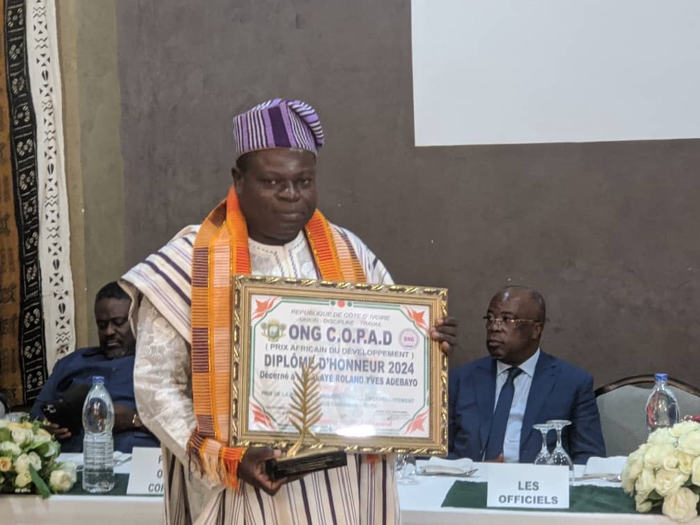 Un homme en tenue traditionnelle tient un diplôme encadré et un trophée avec des personnes assises derrière lui à une table.