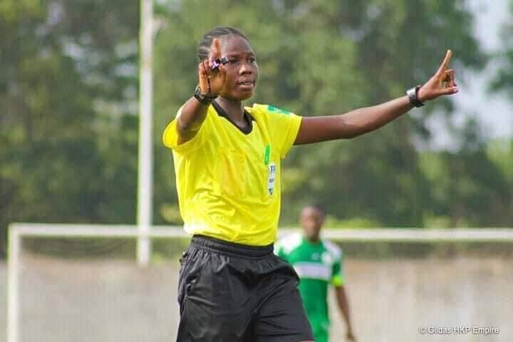 Une arbitre en tenue jaune signale avec les bras levés sur un terrain de football avec un joueur flou en arrière-plan