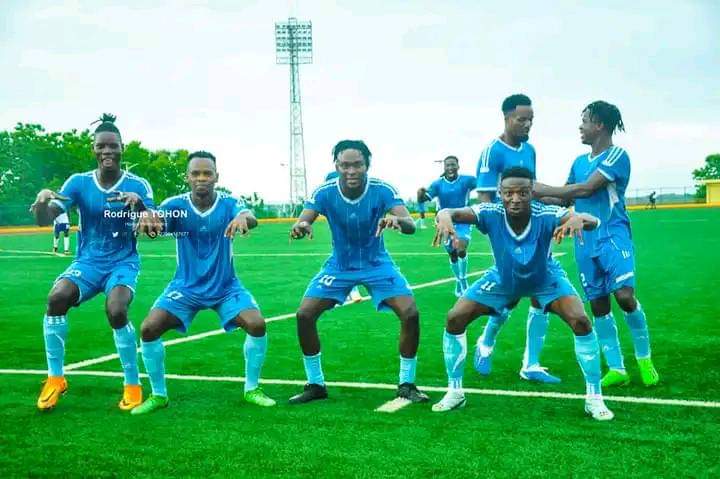 Six joueurs en tenue bleue posant en groupe sur un terrain de football vert sous un ciel nuageux