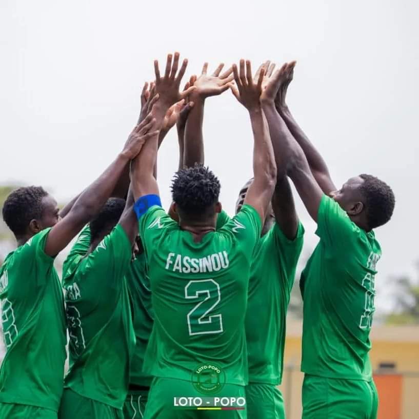 Cinq joueurs en maillots verts, bras levés, célébrant ensemble sur un terrain de football en extérieur