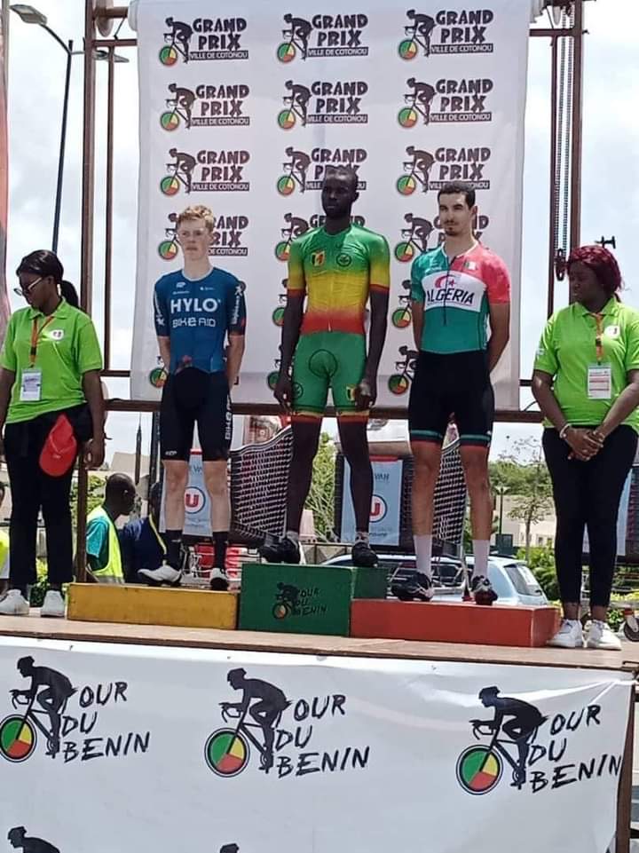 Trois cyclistes en tenue de course debout sur un podium de récompense encadrés par deux femmes en tenue verte