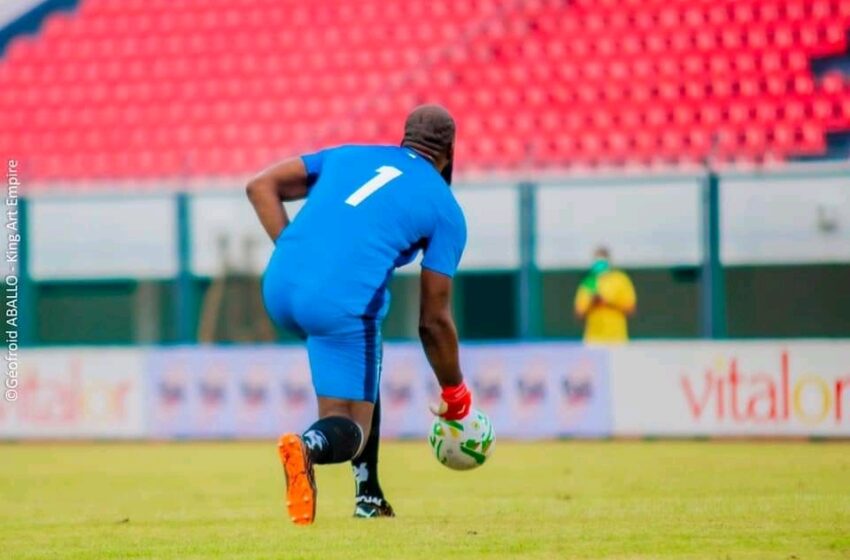 Un joueur de football en tenue bleue s'apprête à lancer un ballon sur un terrain vide avec des gradins rouges en arrière-plan
