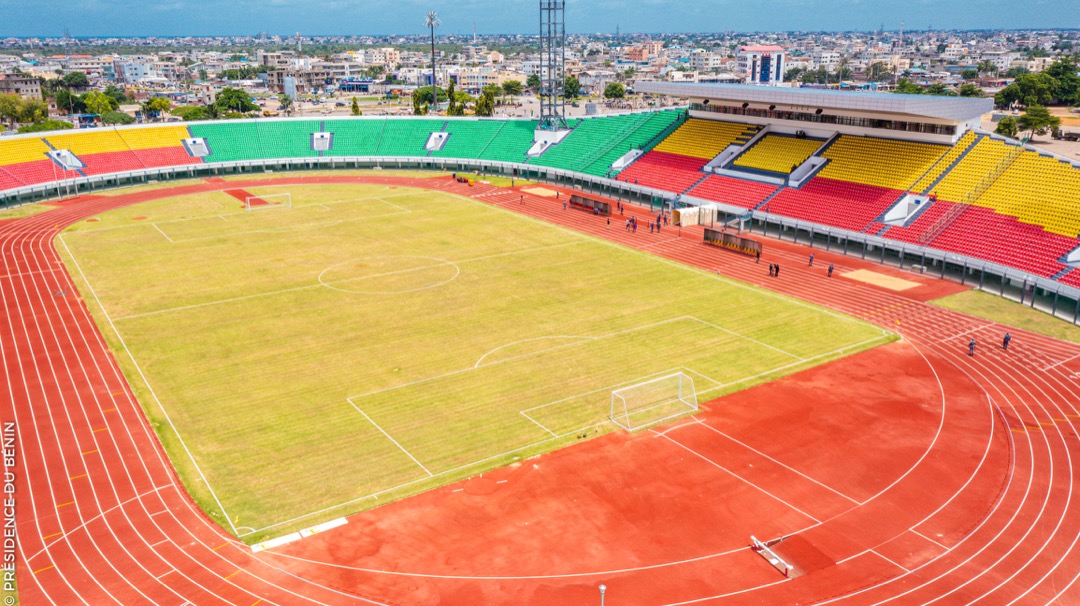 Stade avec pelouse partiellement verte et gradins aux couleurs vives entourés d’une piste d’athlétisme rouge et vue urbaine en arrière-plan