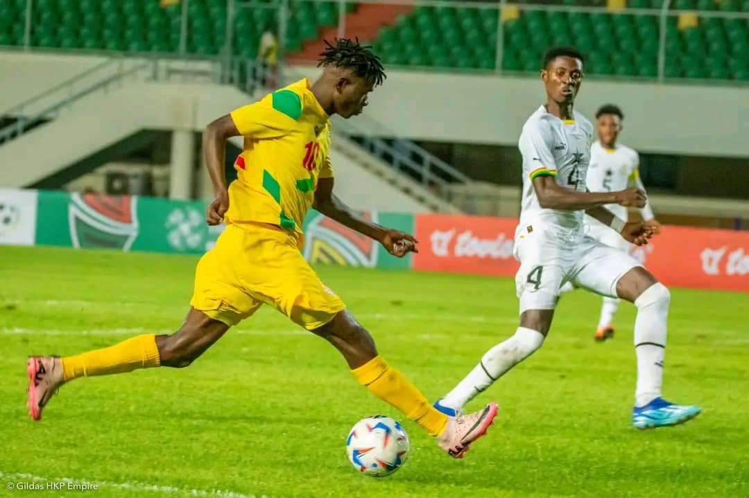 Un joueur en jaune tire au but face à deux défenseurs en blanc sur un terrain de football vert