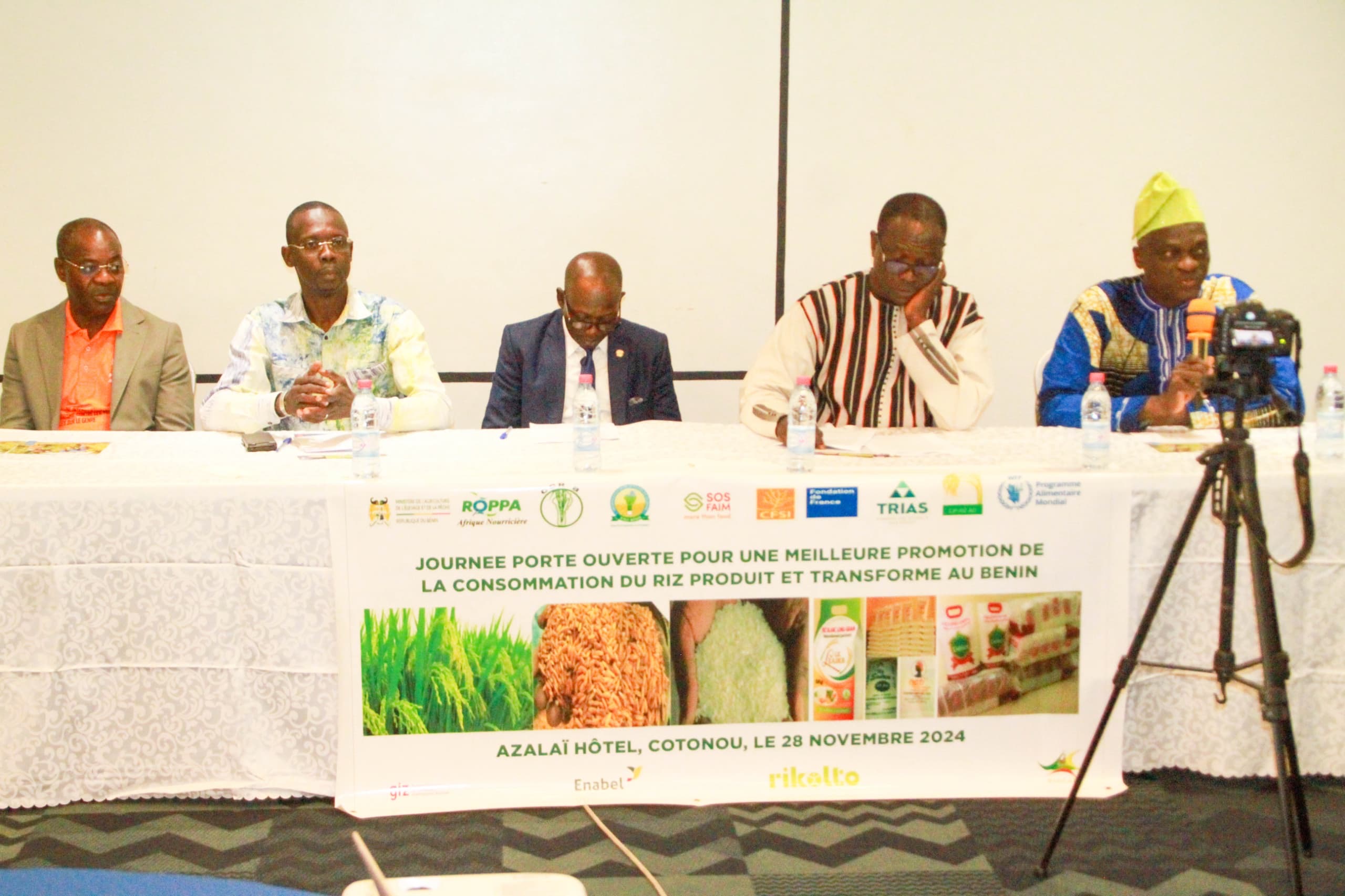 Cinq hommes assis derrière une table avec une bannière sur la promotion de la consommation du riz au Bénin devant un mur blanc et un trépied caméra