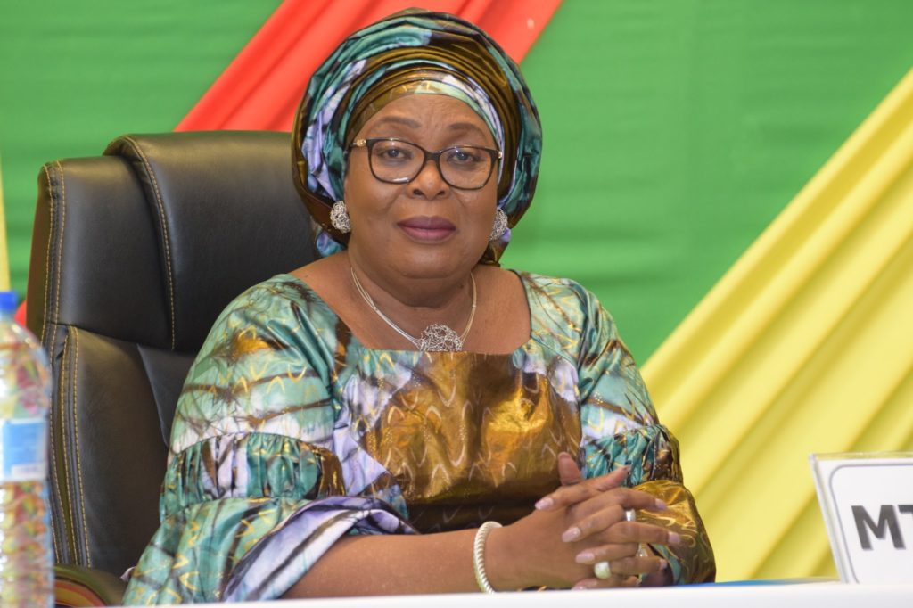 Femme portant un pagne coloré et un foulard assise devant un drapeau aux couleurs vives avec les mains croisées sur une table