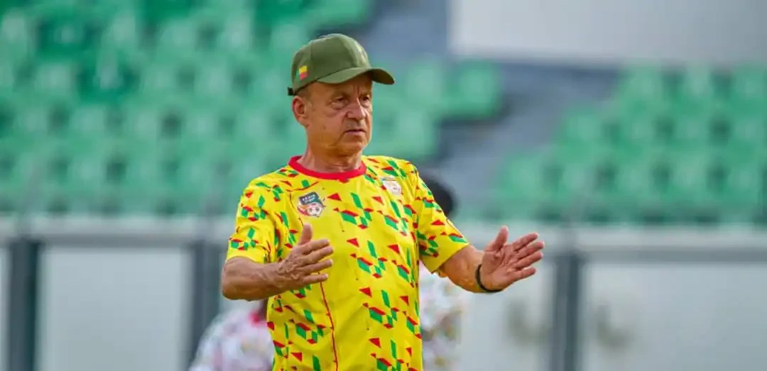 Un homme en tenue sportive jaune et casquette verte fait un geste avec ses mains dans un stade aux gradins verts vides